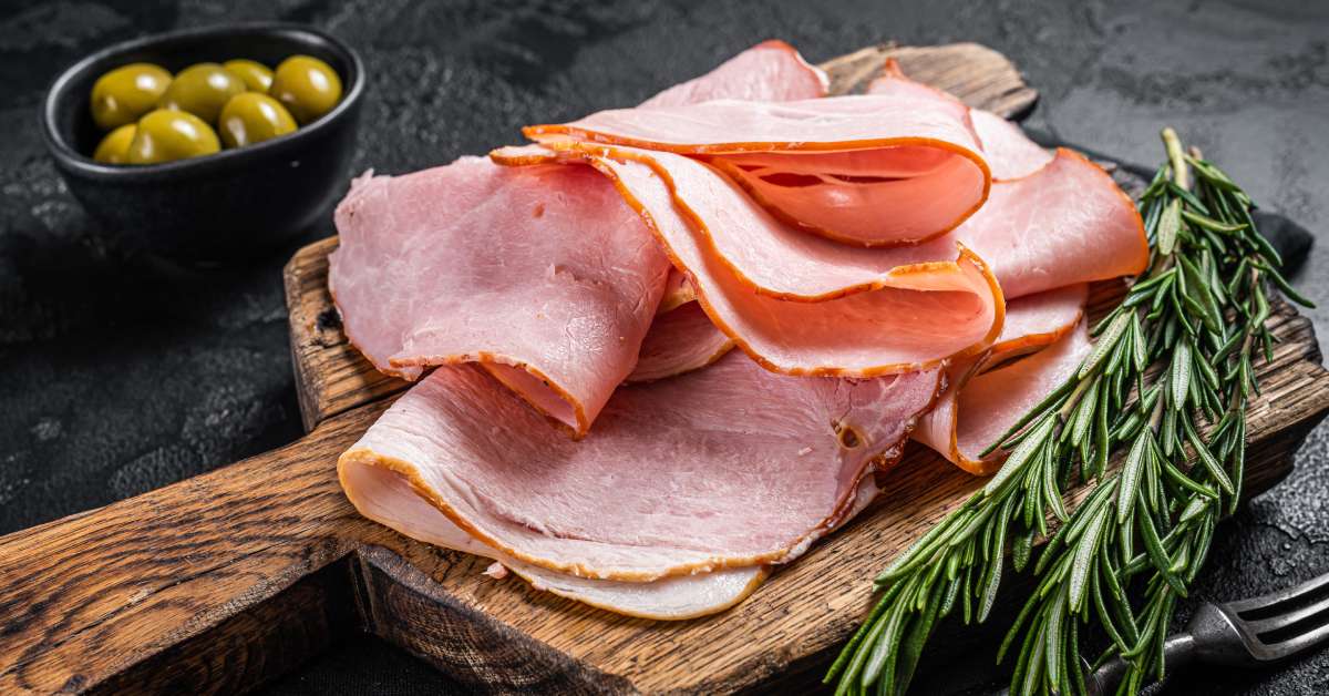 Thin slices of ham piled onto a wooden serving board. Sprigs of rosemary and a small bowl of green olives on either side.