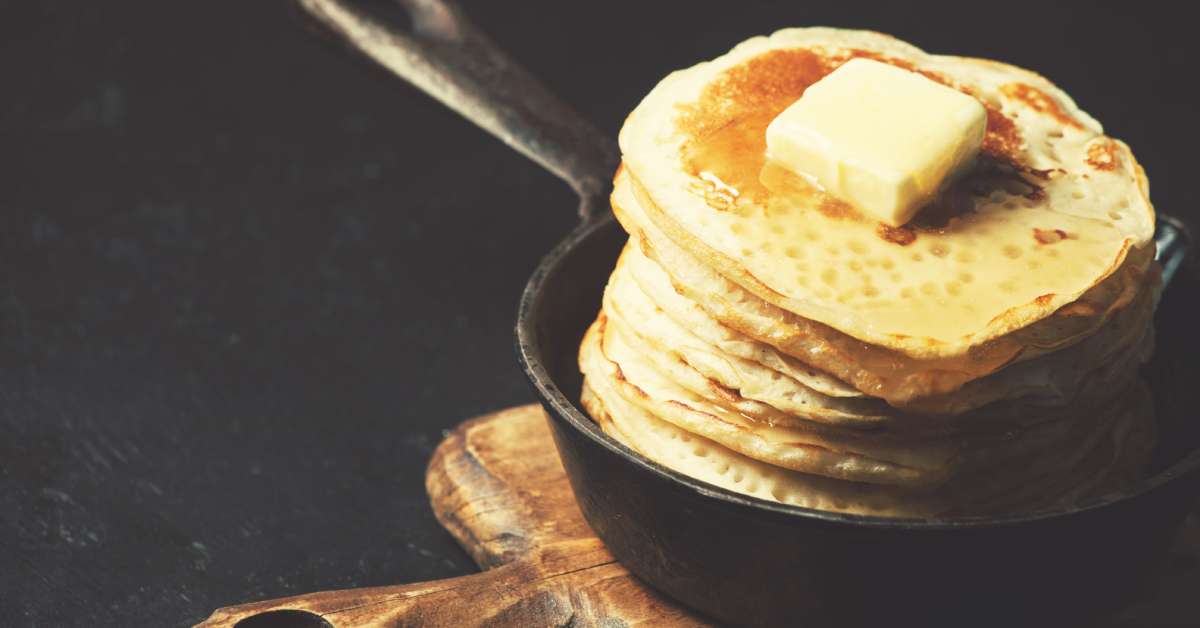 A tall stack of fluffy pancakes with a pad of butter on top sits in a cast iron skillet that rests on a wooden serving tray.